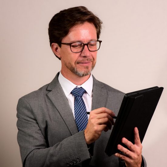 Fabiano Cardozo Homem de terno cinza e gravata azul, usando óculos e interagindo com um tablet enquanto sorri levemente.
