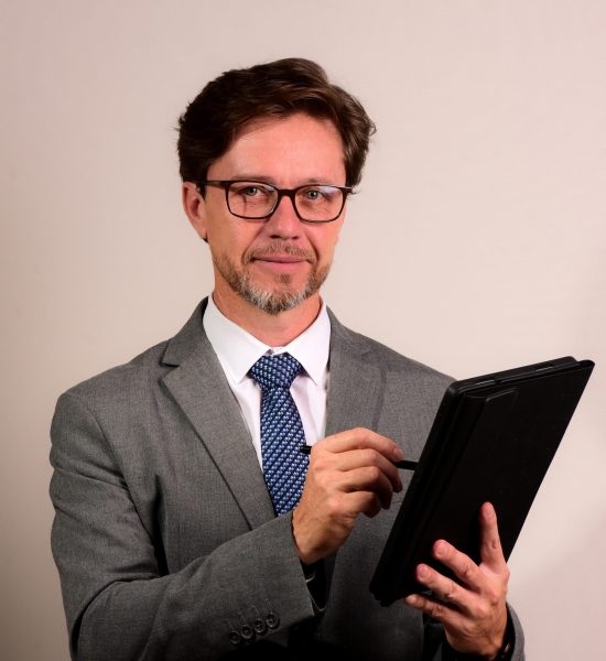 Fabiano Cardozo Homem elegante de óculos, vestindo terno cinza, camisa branca e gravata azul com padrão discreto. Ele segura um tablet preto com uma caneta digital, demonstrando concentração e sofisticação, em um fundo minimalista e neutro.